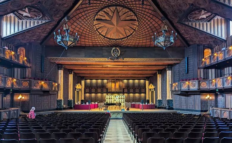 An empty wood-paneled concert hall with a stage, piano, and rows of seats.